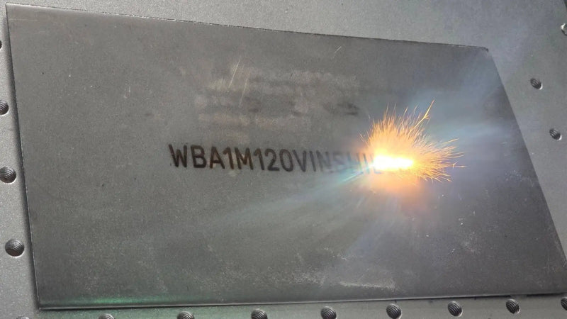 A dark gray metallic plate with engraved alphanumeric text and a bright spark of welding activity.
